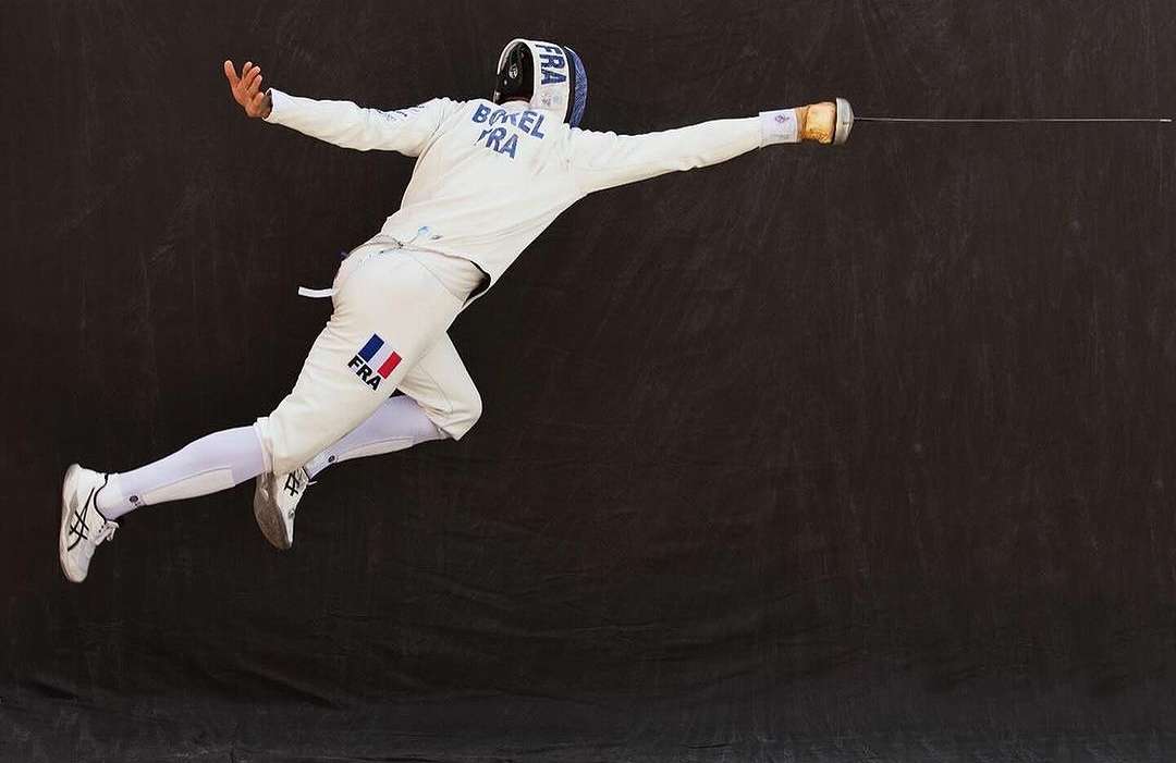Male fencer in mid-air executing a move, wearing white protective gear with the French flag, against a dark background.