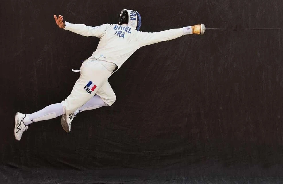 Male fencer in mid-air executing a move, wearing white protective gear with the French flag, against a dark background.