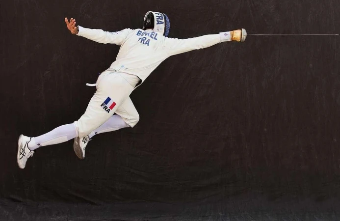 Male fencer in mid-air executing a move, wearing white protective gear with the French flag, against a dark background.
