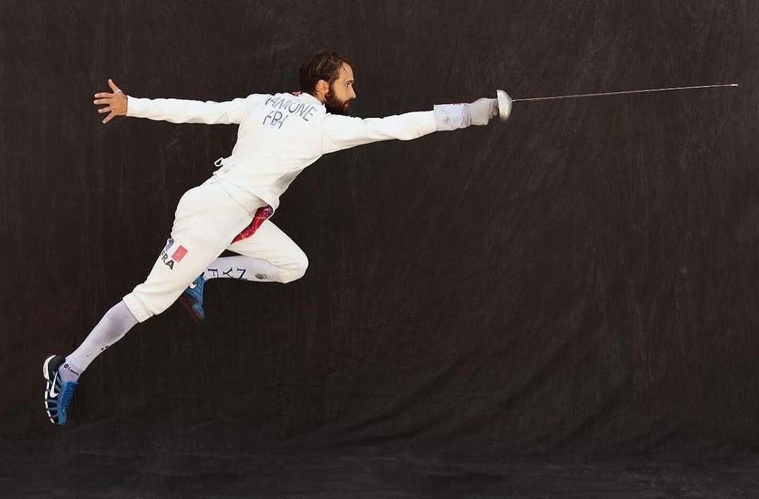 A man in fencing gear in mid-air, lunging forward with a foil, isolated against a dark backdrop, striking a dynamic pose in a sports context.