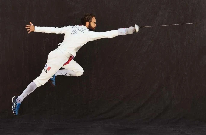 A man in fencing gear in mid-air, lunging forward with a foil, isolated against a dark backdrop, striking a dynamic pose in a sports context.