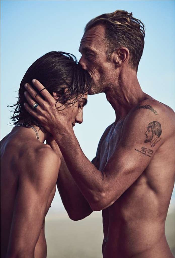 An older adult male kisses the forehead of a younger adult male in an outdoor setting with a blue sky. The subjects are shirtless.