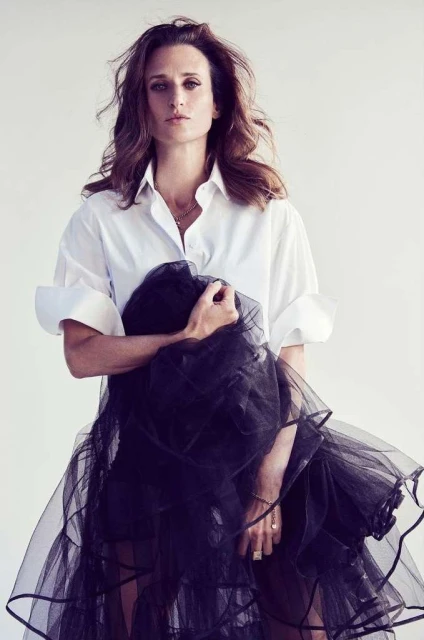 Adult woman in a white button-down shirt holds a black tulle skirt, standing in front of a plain background in a studio shot.
