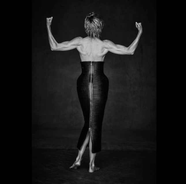 Striking black and white portrait of an adult woman from behind, showcasing toned arms, an elegant leather skirt, and a dimly lit studio setting.