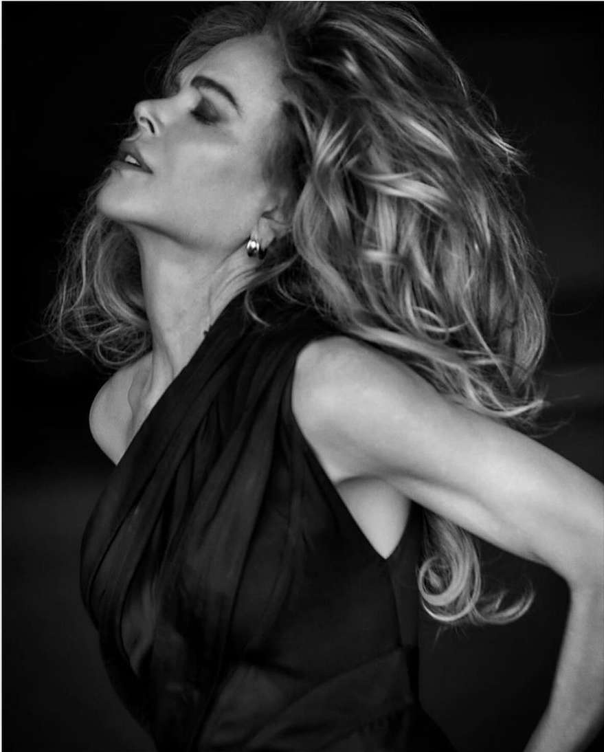 Monochrome portrait of a woman with long wavy hair wearing a black draped dress in a studio setting, looking upward with closed eyes.