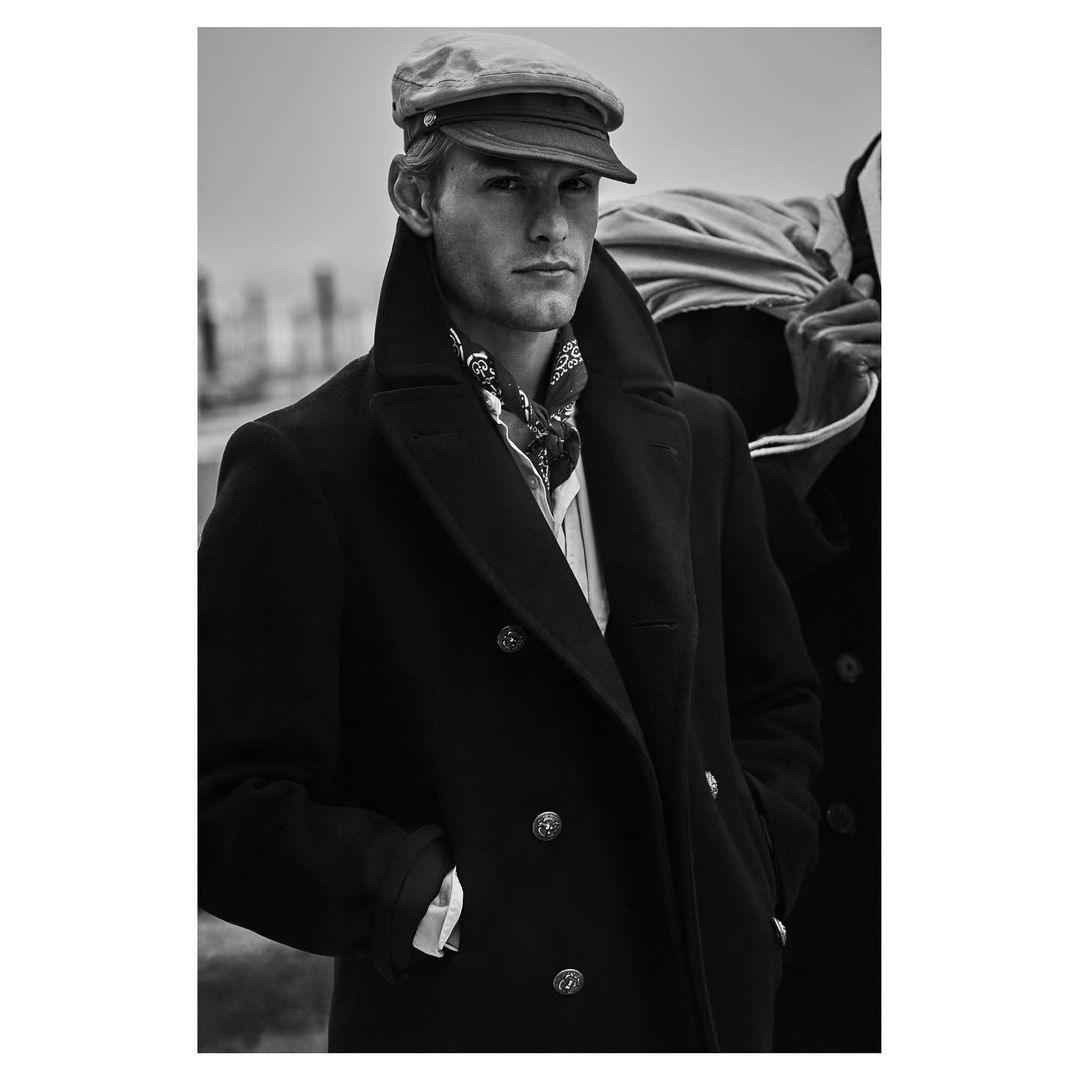 Black-and-white portrait of a male model in a black coat and cap, with a blurred figure in the background on an overcast day.