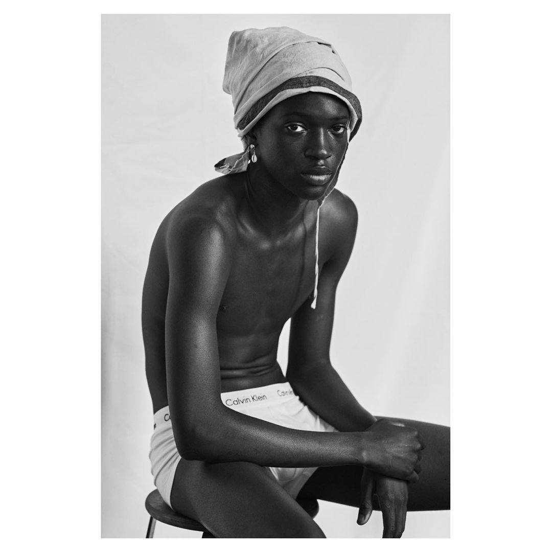 Black and white portrait of a young shirtless adult sitting on a stool with a wrapped head covering, wearing briefs, and facing the camera.