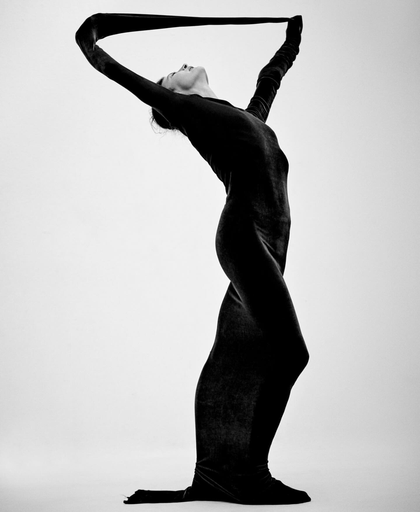 Artistic black-and-white photo of a woman in a full-body dark garment, arching back, and holding fabric overhead in a light studio setting.