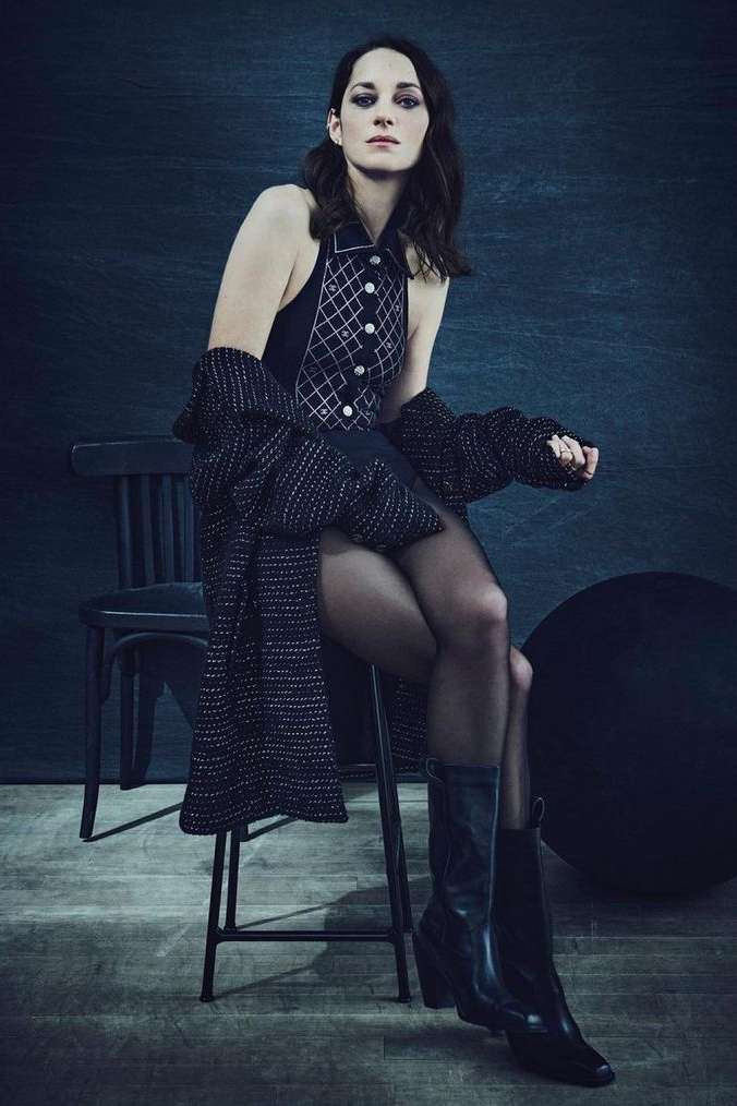 Elegant woman in a patterned dress and boots, seated on a stool in a moody studio setting, exuding style and sophistication.