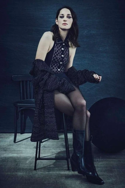 Elegant woman in a patterned dress and boots, seated on a stool in a moody studio setting, exuding style and sophistication.