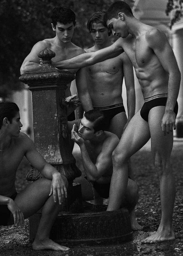 Black-and-white group portrait of five male fashion models near an ornate water fountain in an outdoor setting, minimalist style.
