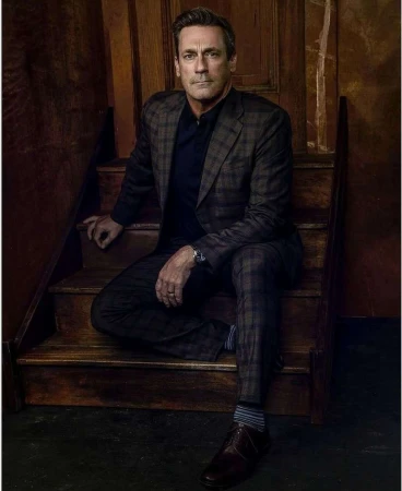 Jon Hamm seated on wooden stairs wearing a plaid blazer and dark trousers by Matthew Brookes