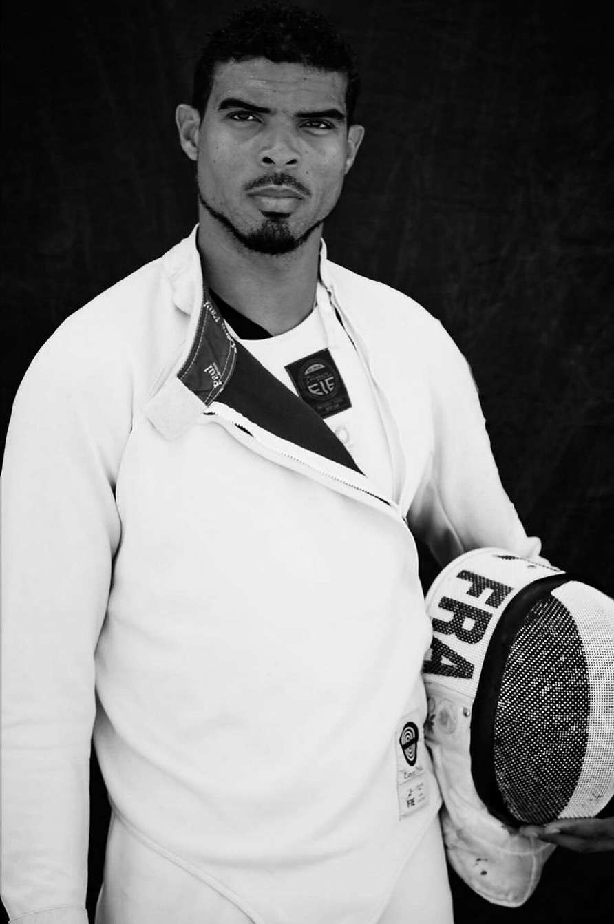 Monochrome portrait of a male fencer in uniform holding a mask, against a dark background. The image highlights sportsmanship.