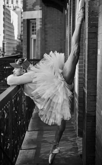 Black and white portrait of a ballet dancer, wearing a tutu and pointe shoes, doing a split against a brick wall on a balcony.