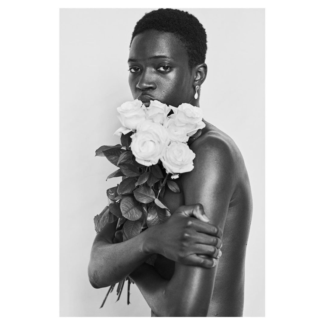 Striking black and white portrait of a young African male model with a bouquet of white roses, set against a plain background. Minimalist beauty.