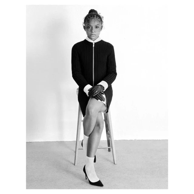 Black and white portrait of a woman in a dress with a veil, seated on a stool. She's wearing heels, and the background is a plain white wall.
