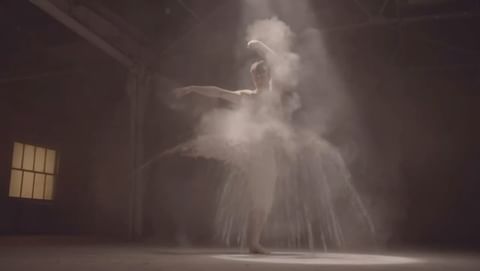 Ballerina performs in a cloud of white powder against a dark background with a single window and overhead stage lighting.