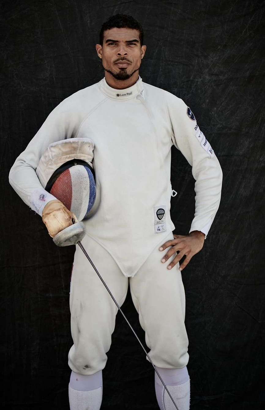 A male fencer in white uniform poses with his foil and mask against a dark background in a studio setting. He has a serious expression.