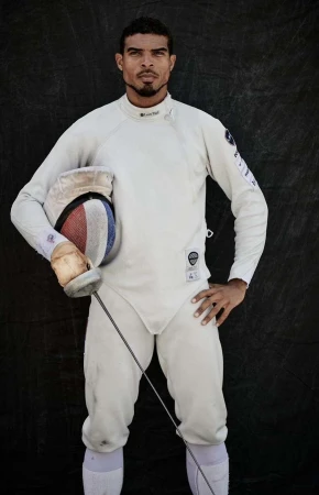 A male fencer in white uniform poses with his foil and mask against a dark background in a studio setting. He has a serious expression.