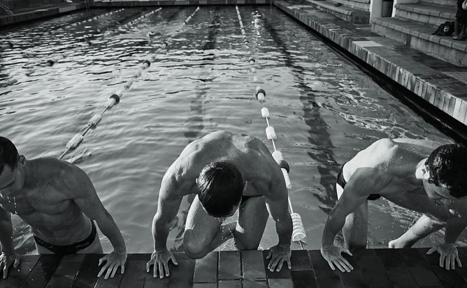 Three muscular swimmers emerge from a pool, grasping the tiled edge, water glistening in black and white. Lanes are visible behind them.