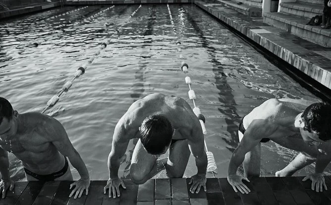 Three muscular swimmers emerge from a pool, grasping the tiled edge, water glistening in black and white. Lanes are visible behind them.