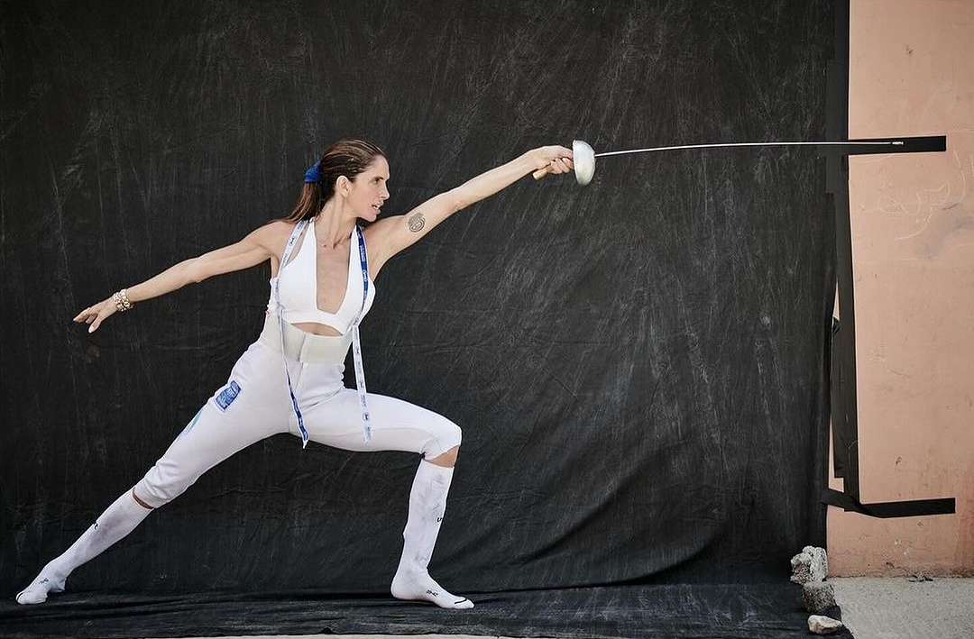 Female fencer in white uniform lunges forward with foil against a textured black backdrop with a pink wall in the background.