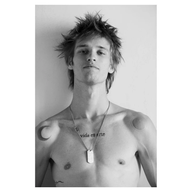 Black and white portrait of a young adult male with tousled hair, tattoos, and a military-style necklace, posing in front of a plain wall.