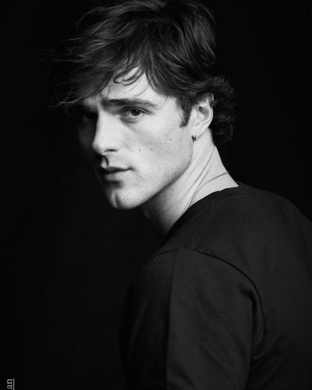 Monochrome portrait of a young adult male with medium-length brown hair, dark t-shirt, and silver chain against a black background.