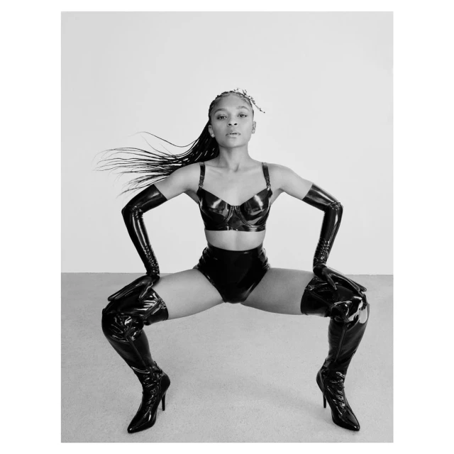 Striking portrait of a woman in black latex bra top, high-waisted briefs, gloves, and thigh-high boots, posing against a neutral backdrop.
