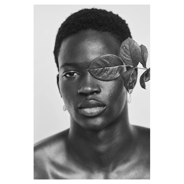 Monochrome portrait of a Black man with a leaf partially obscuring his face, set against a neutral background in a studio environment.