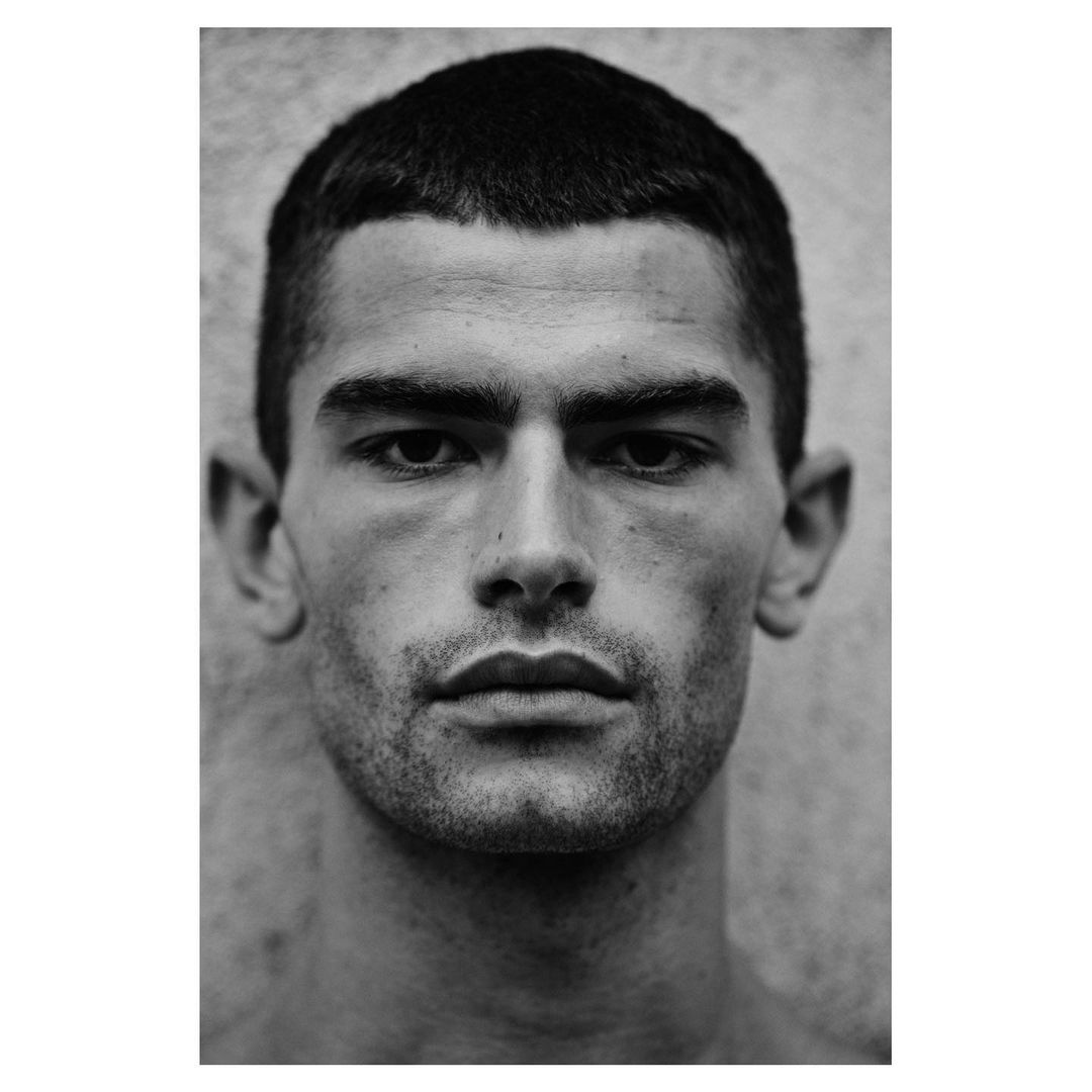 Monochrome portrait of an adult male with short dark hair and stubble, facing forward against a textured grey background, studio lighting.