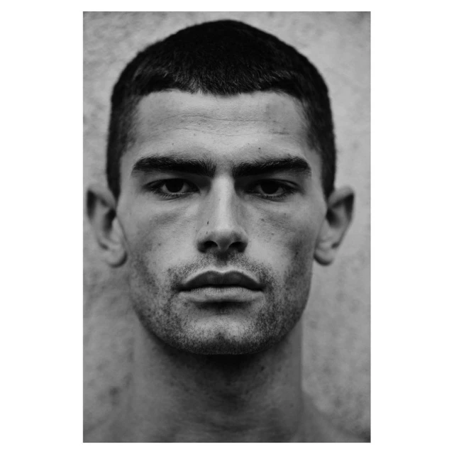 Monochrome portrait of an adult male with short dark hair and stubble, facing forward against a textured grey background, studio lighting.