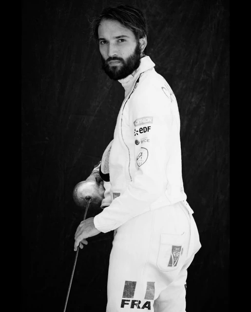 Monochrome portrait of a male fencer in a white uniform with a sword, set against a dark background, captured in a studio setting.