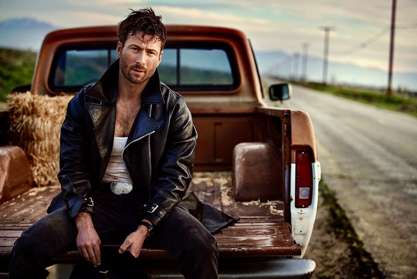 Adult male in leather jacket poses on the back of a vintage pickup truck against a rural road and horizon at golden hour.