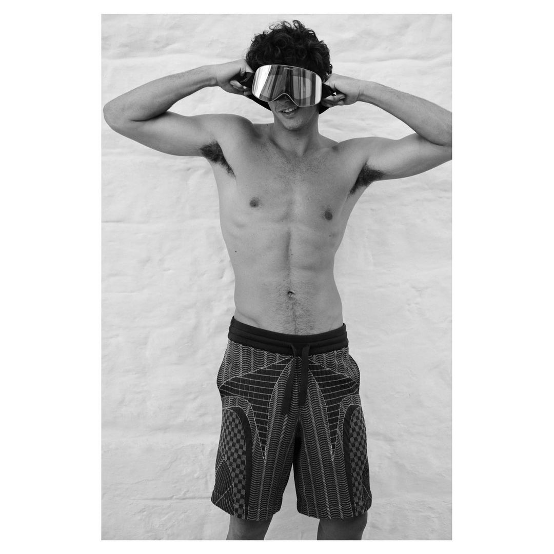 Black and white shot of a shirtless man with dark curly hair and goggles on his forehead, wearing patterned shorts against a textured white wall.