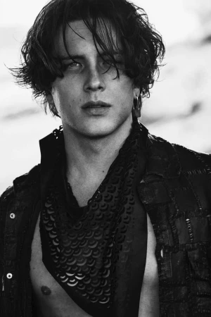 Monochrome portrait of a young man with dark tousled hair, wearing a patterned patterned scarf and dark jacket.
