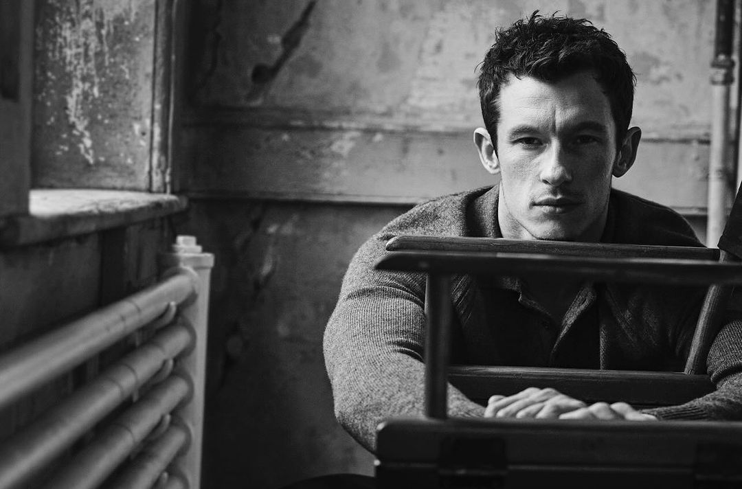 Black and white portrait of a pensive adult man in a grey sweater, leaning forward in an indoor setting with textured walls and window frame.
