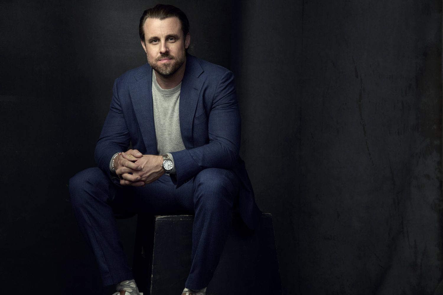 Man in a blue suit and grey t-shirt seated against a dark textured studio backdrop