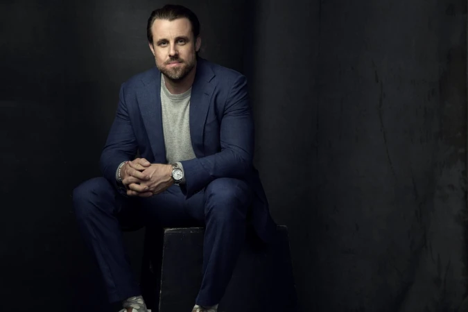 Man in a blue suit and grey t-shirt seated against a dark textured studio backdrop