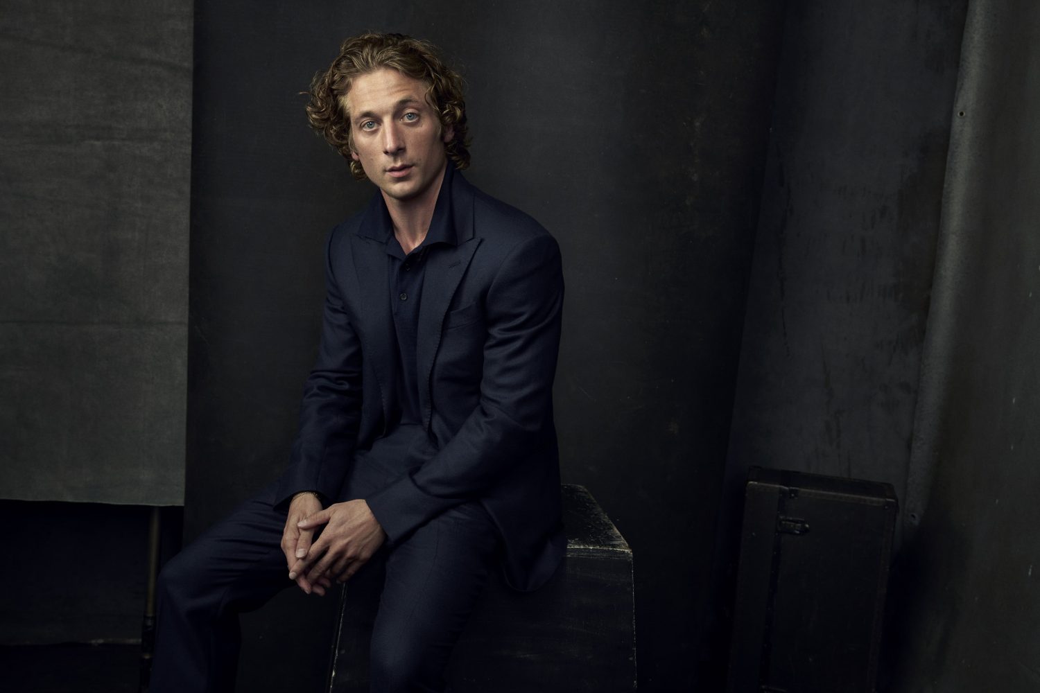 Indoor portrait of an adult male with curly brown hair, wearing a navy suit and sitting on a rustic wooden box against a dark background.