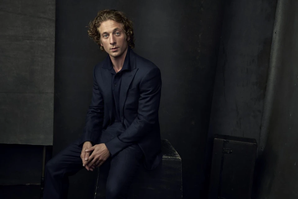 Indoor portrait of an adult male with curly brown hair, wearing a navy suit and sitting on a rustic wooden box against a dark background.