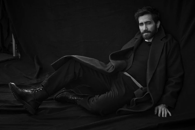 Jake Gyllenhaal seated on a dark draped floor wearing a heavy black coat, dark trousers, and leather boots.