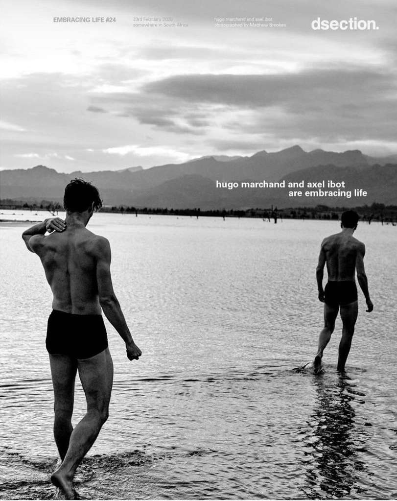 Two shirtless men wading in shallow water towards mountains under cloudy sky, black and white photograph. Minimal clothing, ripples in water.