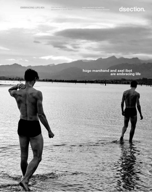 Two shirtless men wading in shallow water towards mountains under cloudy sky, black and white photograph. Minimal clothing, ripples in water.