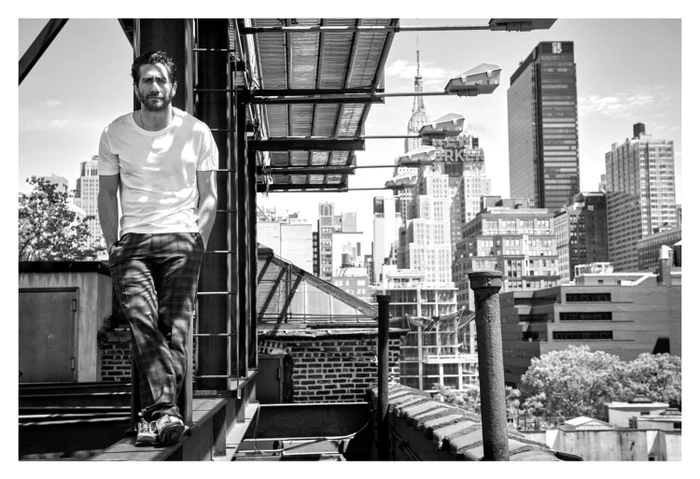 Monochrome portrait of Jake Gyllenhaal leaning against a steel beam on a city rooftop with skyscrapers in the background.