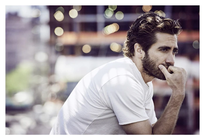 Portrait of Jake Gyllenhaal with a beard wearing a white t-shirt against an out-of-focus city background