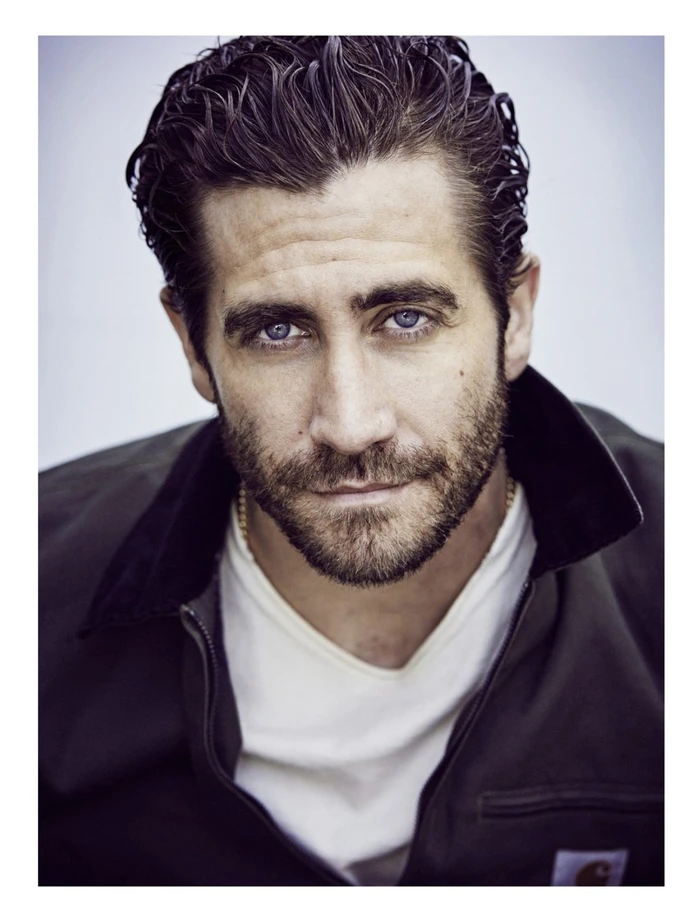 Close-up portrait of Jake Gyllenhaal with dark wet hair wearing a black jacket and white t-shirt against a plain white background