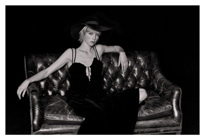 Woman in black velvet dress and wide-brim hat seated on a worn leather chesterfield sofa in a dark studio setting