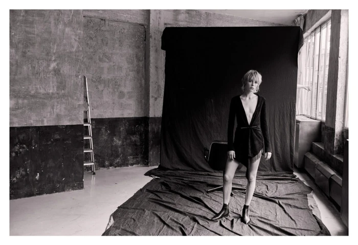 Model in dark wrap dress and boots standing before a black backdrop in a warehouse studio by Matthew Brookes