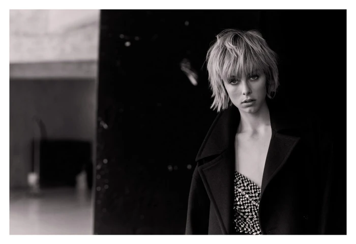 Black and white portrait of a woman with short shaggy hair wearing a heavy coat and a sparkling top against a dark background